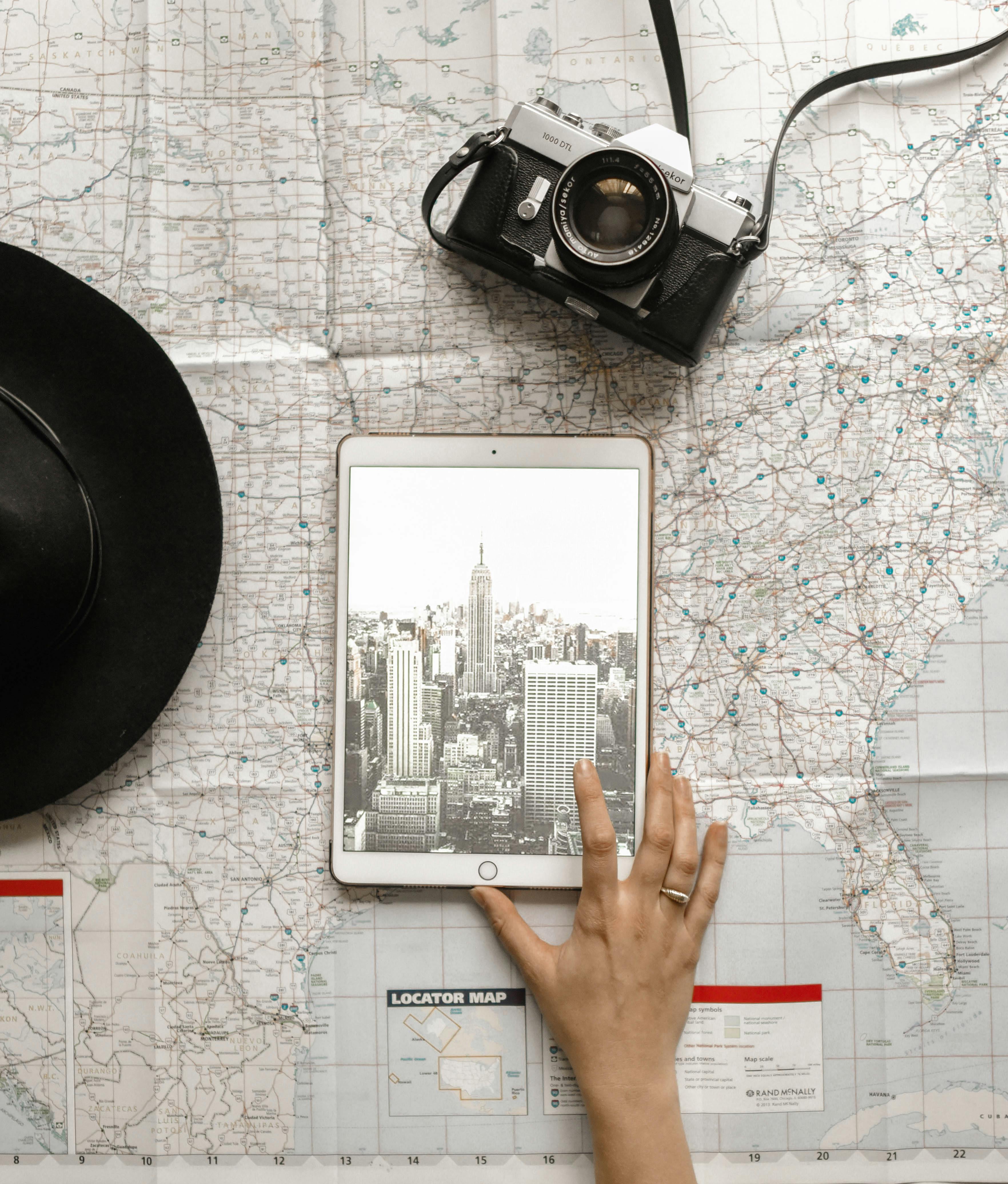 Hand holding a tablet displaying a black and white cityscape on a folded map, with a vintage camera and black hat nearby.
