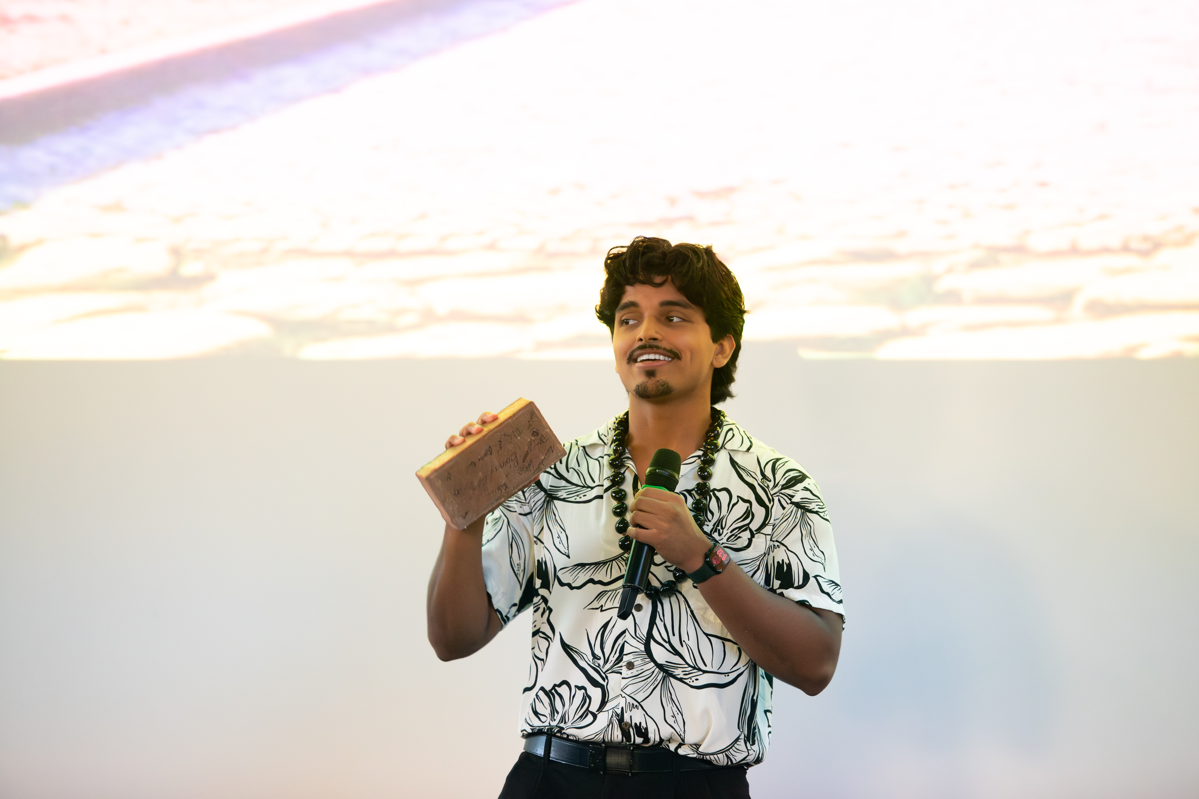 Man in floral shirt holding a microphone and showing a brown rectangular object while speaking.