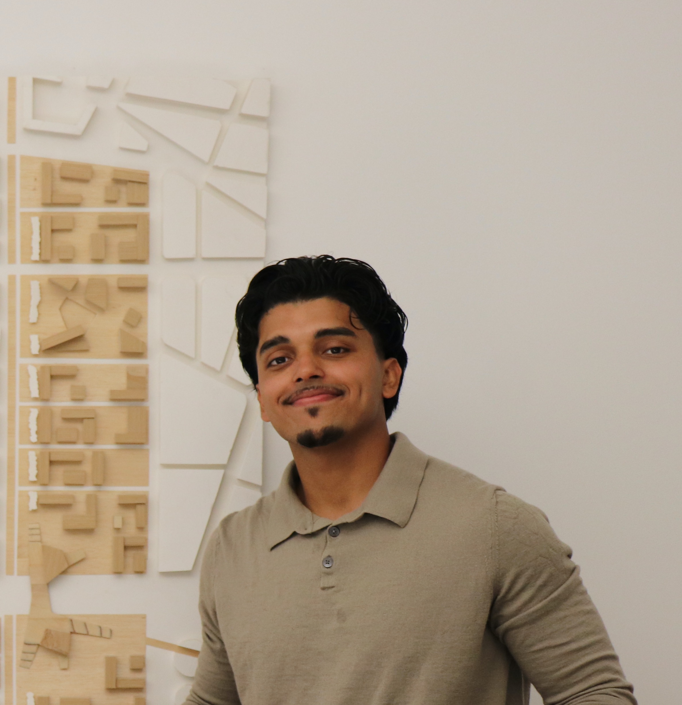 Smiling man with dark hair wearing a beige long-sleeve collared shirt standing in front of a wall with a wooden architectural model.