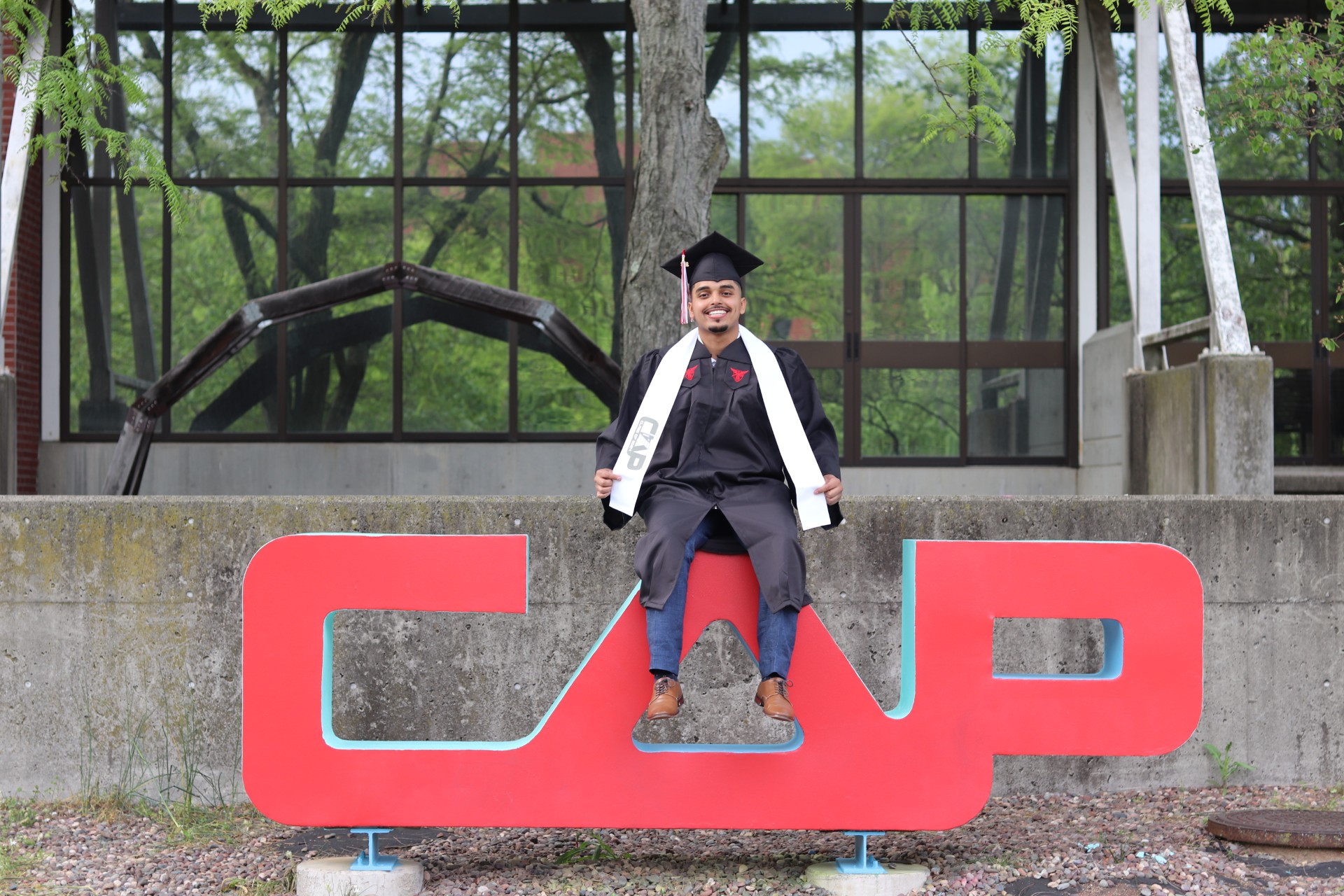 Young man in graduation cap and gown sitting on large red letters 'CAP' outdoors in front of a building with glass windows.