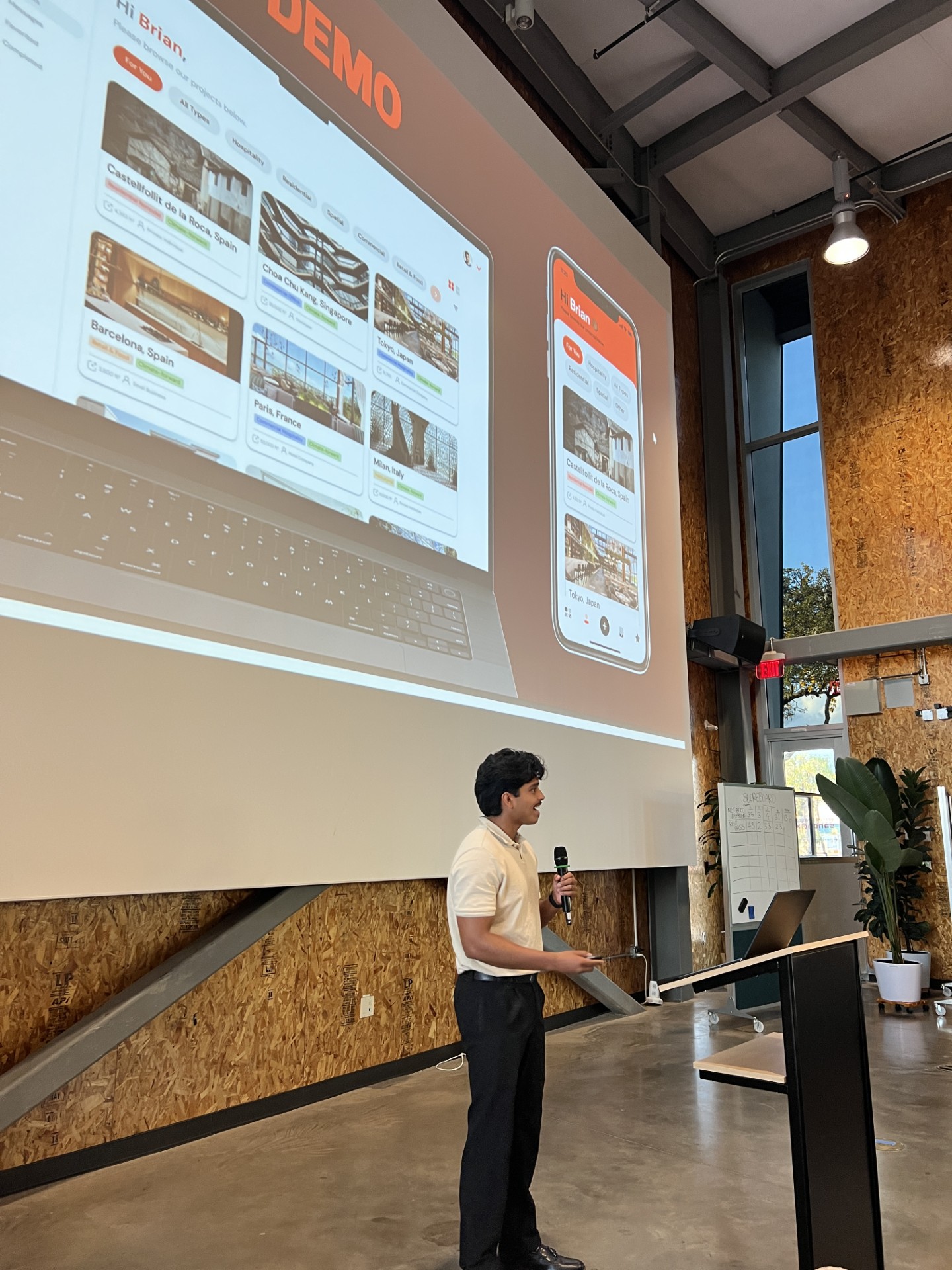 Man in a white shirt holding a microphone and presenting a digital project interface projected on a large screen in an industrial-style room.