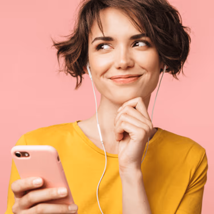 Jeune femme souriante portant un t-shirt jaune, écoutant de la musique avec des écouteurs blancs et tenant un smartphone rose sur un fond rose.
