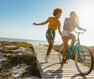Deux femmes souriantes sur une piste en bois près de la mer, l'une poussant un vélo bleu pendant que l'autre court à côté.