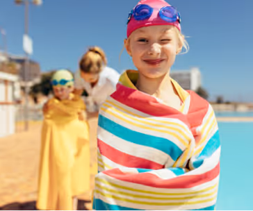 Jeune fille souriante enveloppée dans une serviette rayée colorée au bord d'une piscine, portant un bonnet et des lunettes de bain roses.