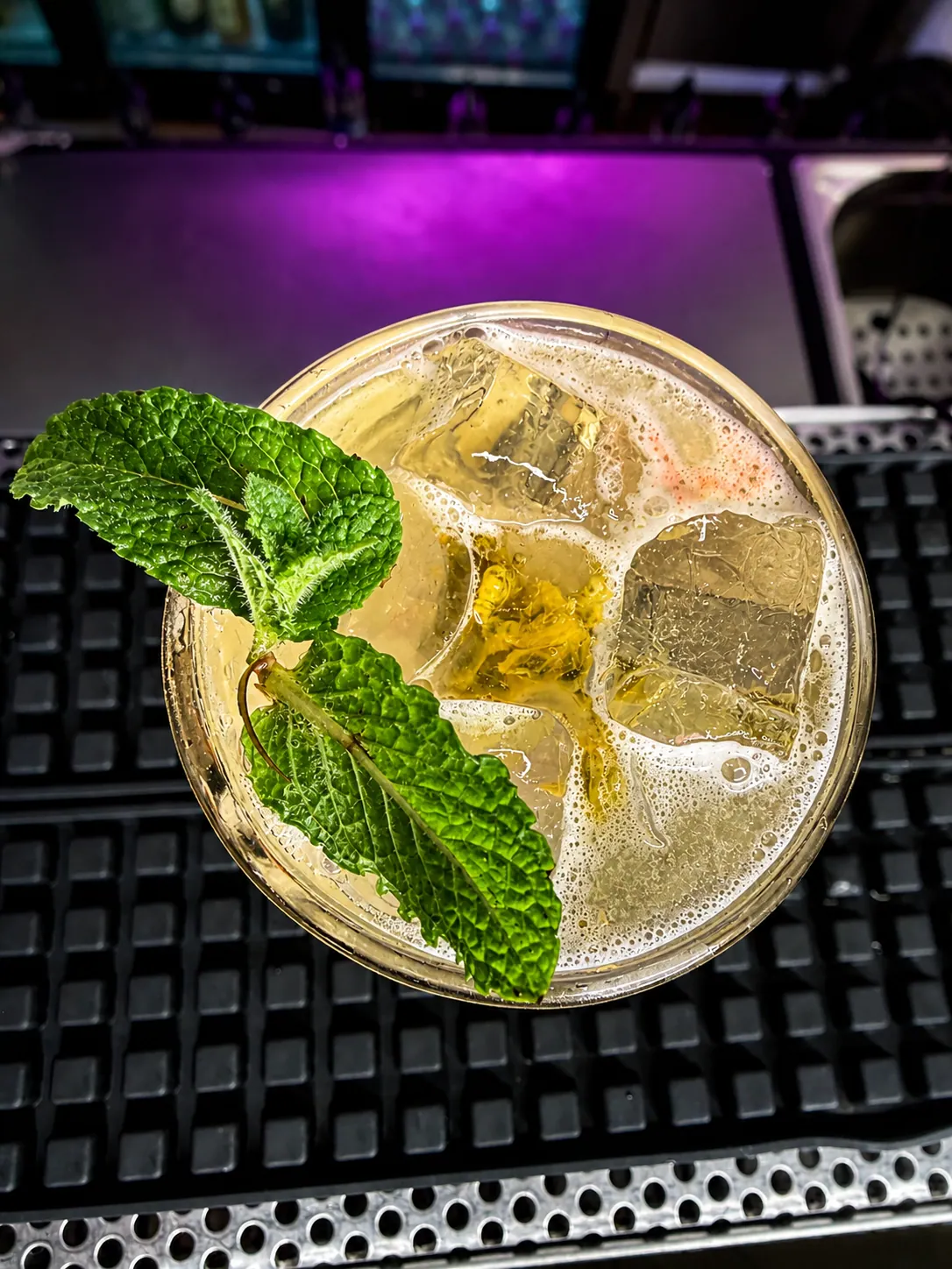 Cocktail con cubetti di ghiaccio e foglie di menta fresca su un bancone da bar con riflessi di luce viola.