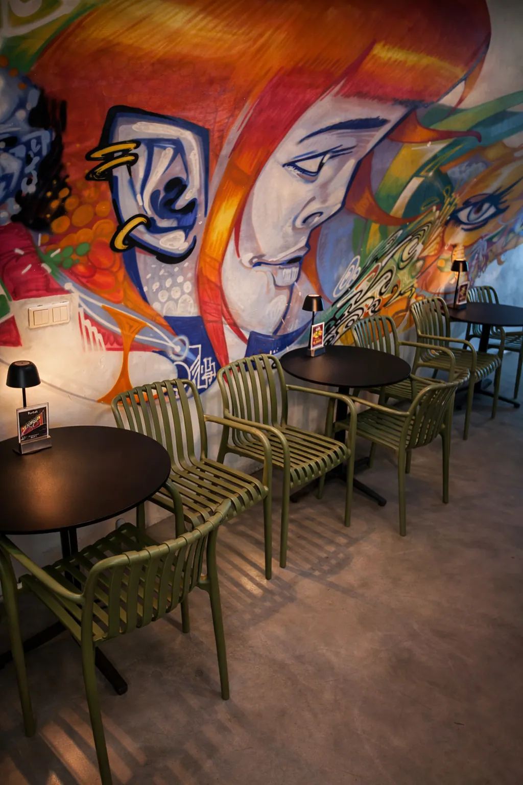 Sedute di un caffè al chiuso con sedie verdi e tavoli neri davanti a un murale colorato con un volto.