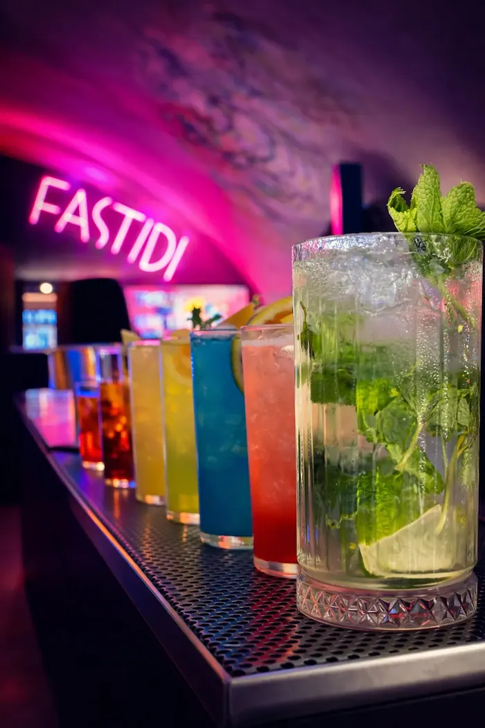 Fila di cocktail colorati allineati su un bancone con un’insegna al neon che dice FASTIDI sullo sfondo.