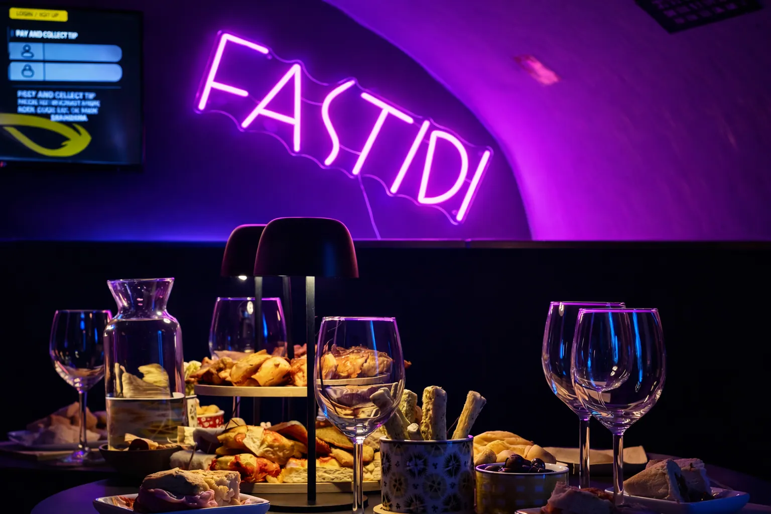 Tavolo con bicchieri, snack e una caraffa d’acqua illuminato da un’insegna al neon viola con la scritta FASTIDI.