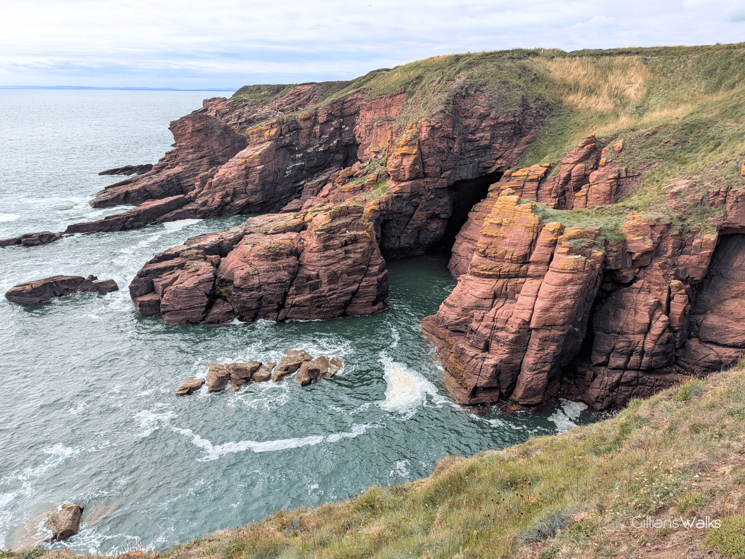 Dramatic red sandstone cliffs with a sea cave meet turquoise ocean waves below.