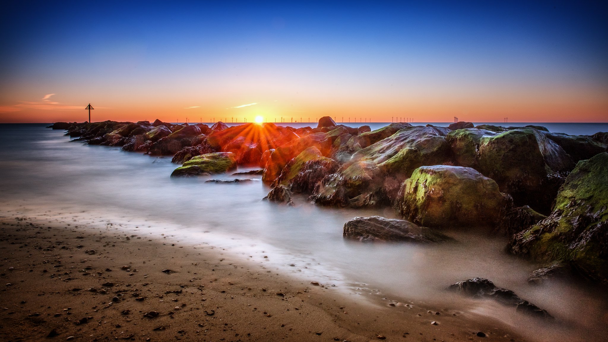 Sunset over rocky beach with waves, golden sky, and distant bridge at dusk