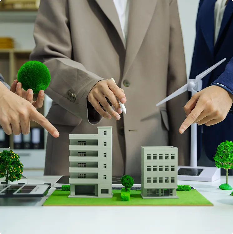 Three people in business attire pointing to a sustainable urban planning model with buildings, greenery, and a wind turbine on a table.