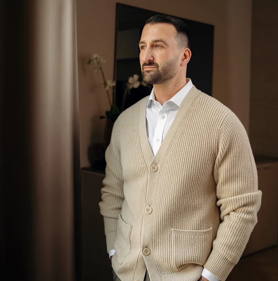Man with short dark hair and beard wearing a beige knit cardigan over a white shirt looking thoughtfully out of a window indoors.