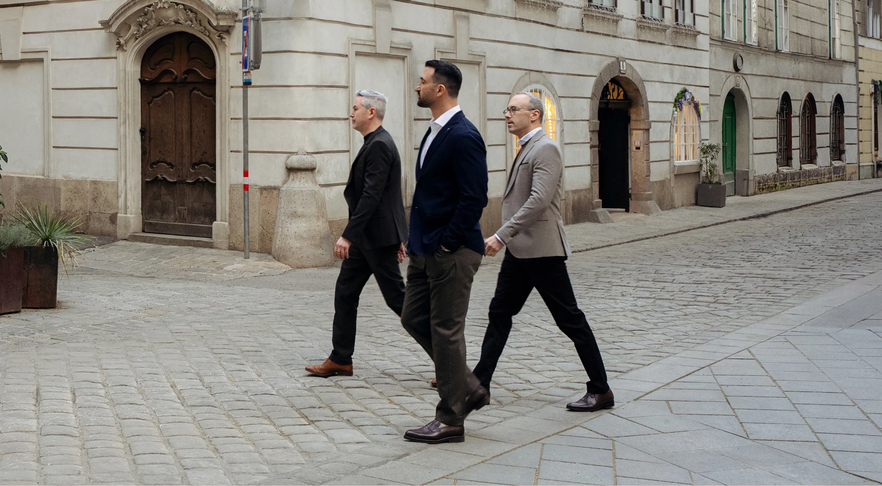 Three men in business attire walking on a cobblestone street in front of historic buildings.