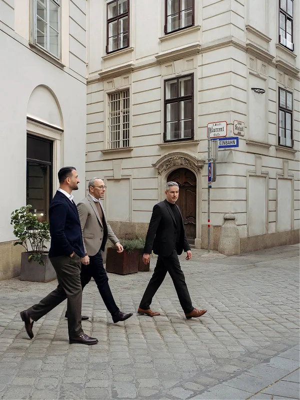 Three men in business attire walking on a cobblestone street in front of a historic building with arched doorway and street signs.