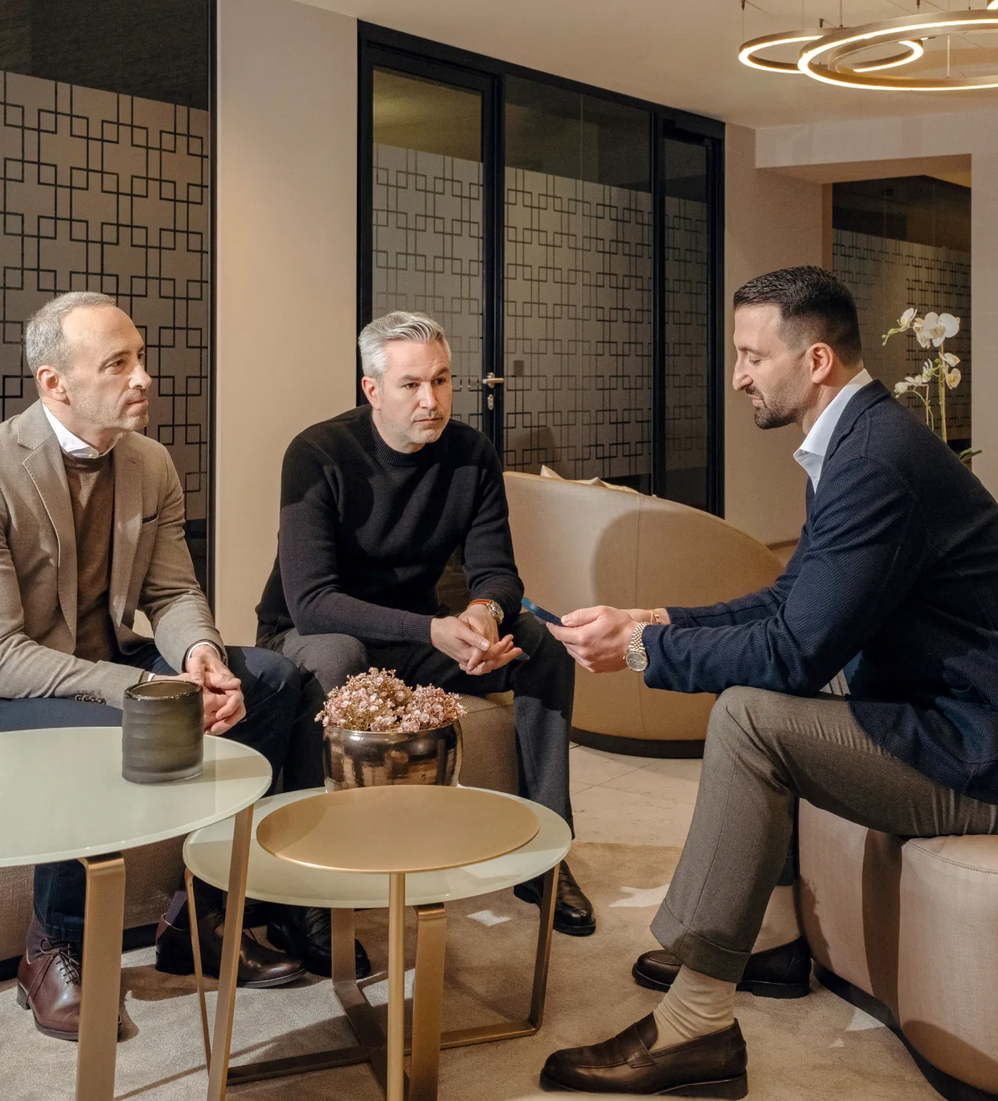 Three businessmen in a modern office lounge engaging in a serious discussion, with two seated on a sofa and one on a chair holding a phone.