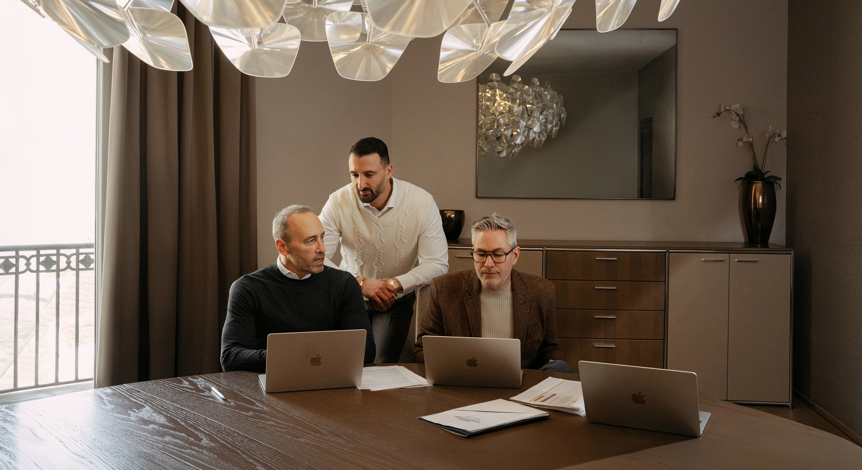 Three men working on laptops around a table with documents in a modern office room.