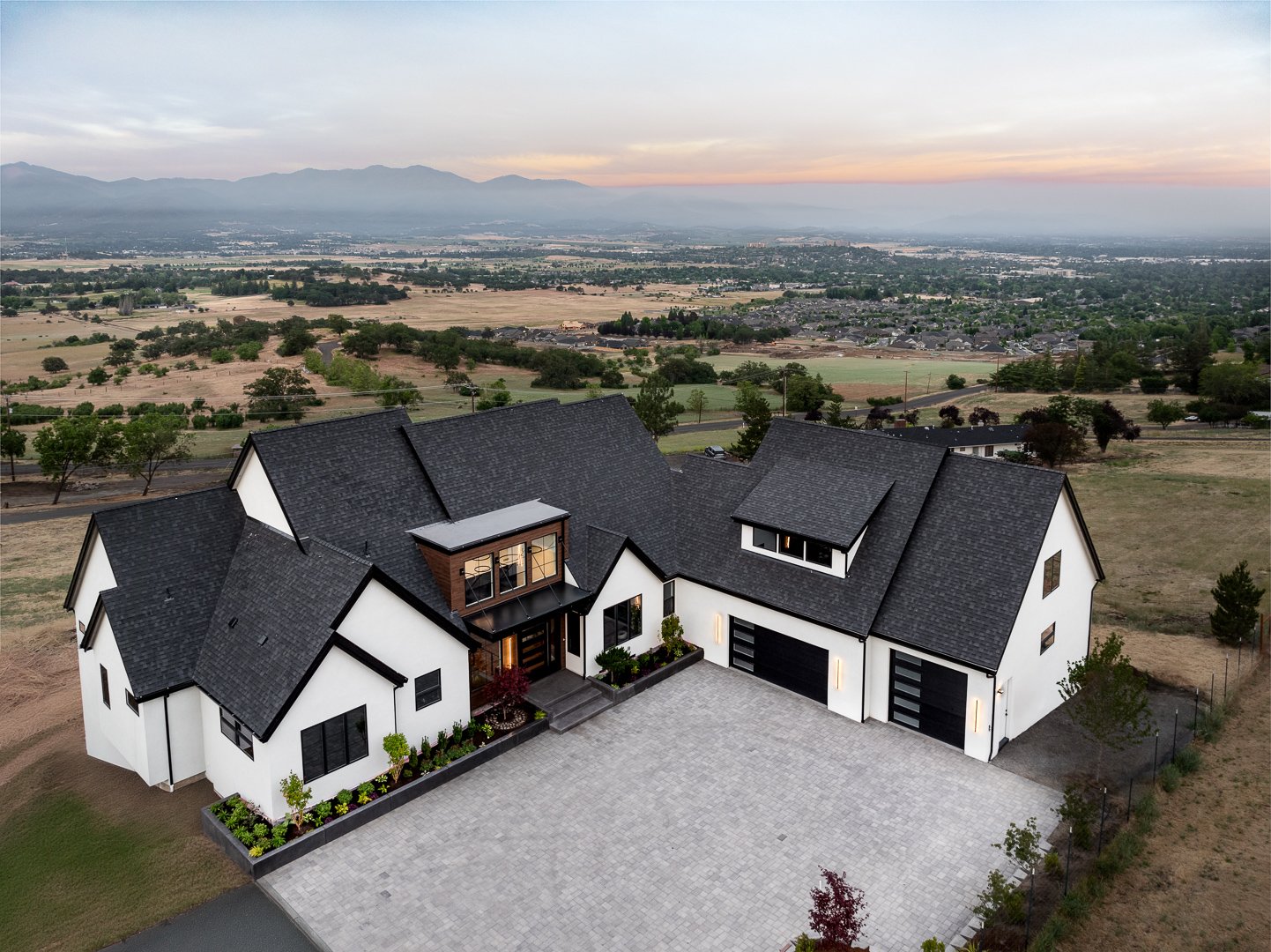 Modern white house with black roof and large paved driveway, surrounded by open fields and distant mountains at sunset.