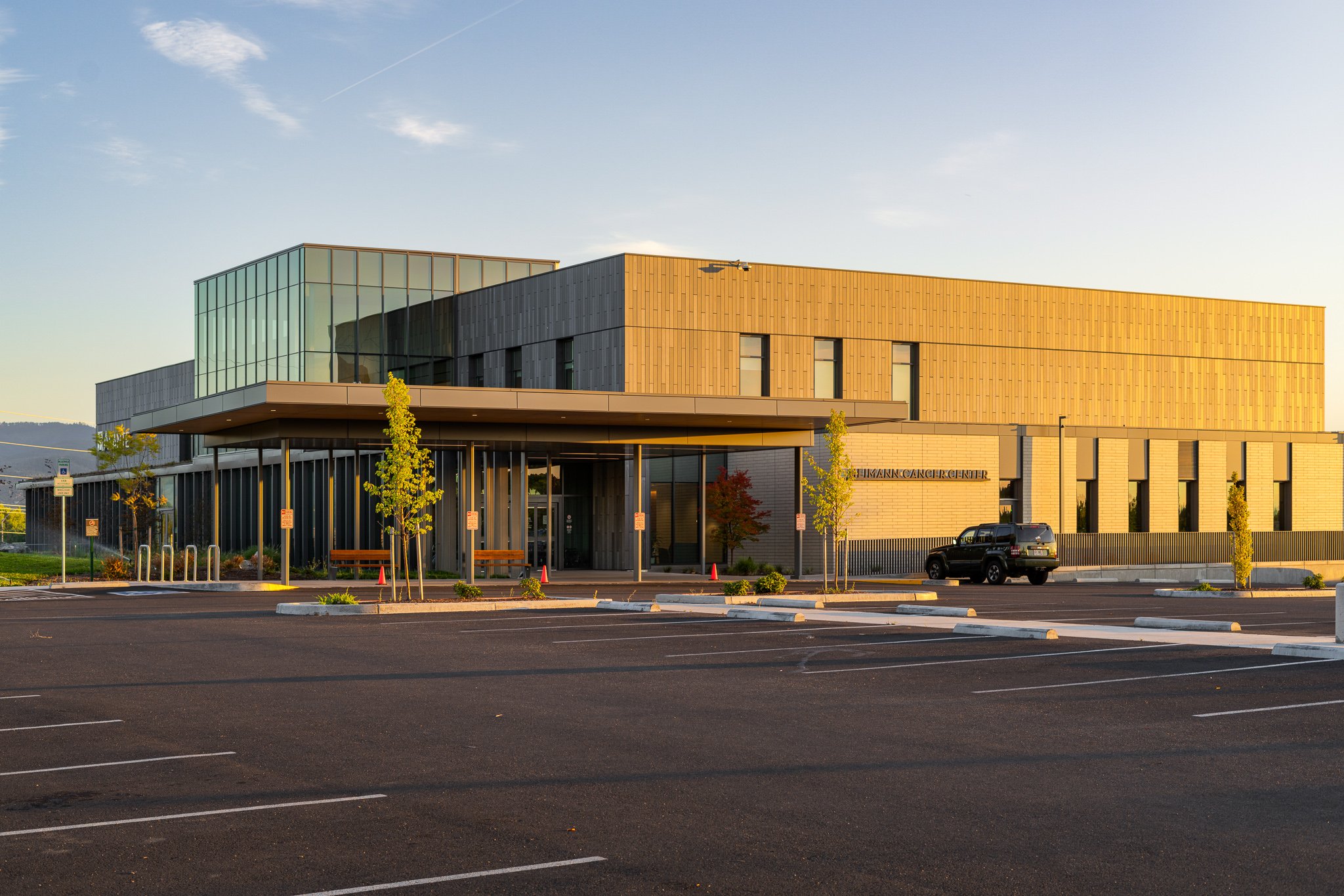Modern cancer center building with large glass windows and empty parking lot in front at sunset.