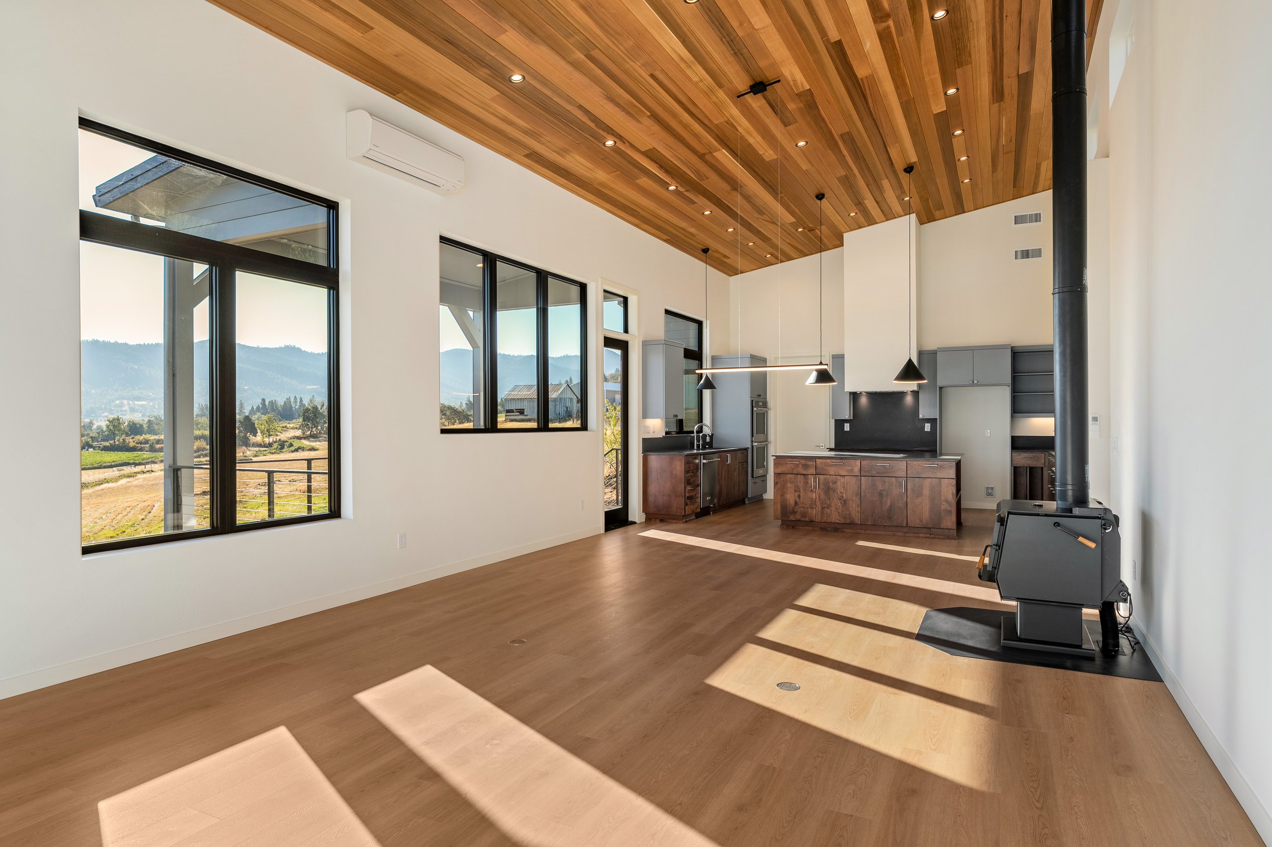 Spacious modern kitchen with wood flooring, wooden ceiling, large windows showing rural landscape, and a black wood stove.