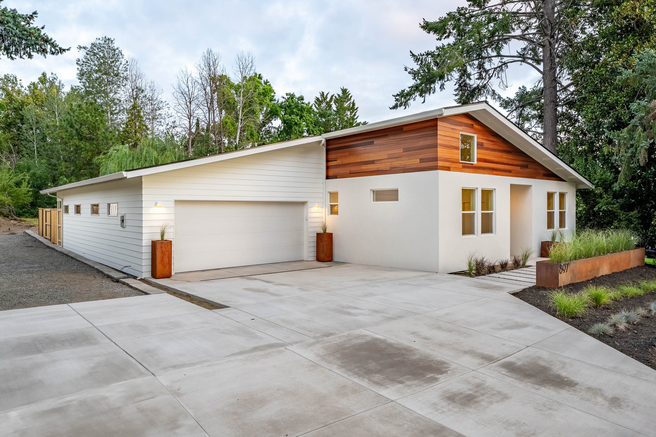 Modern single-story house with white siding, wooden panel accents, large driveway, and surrounding greenery.