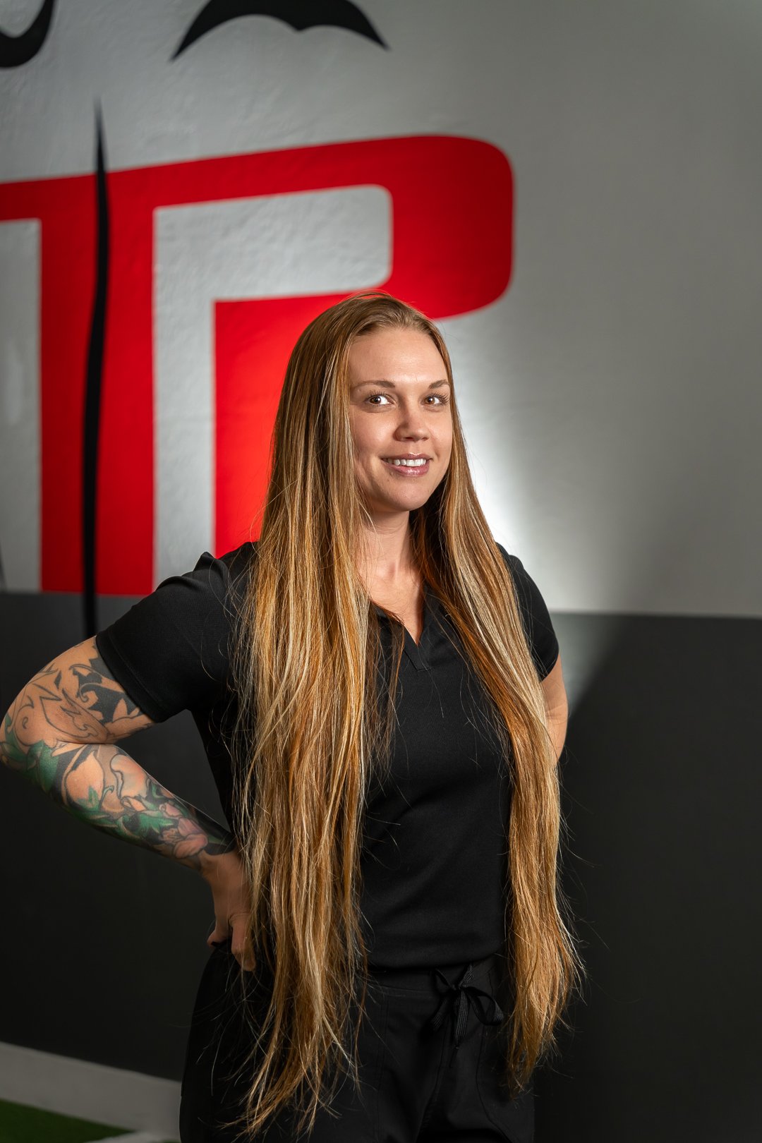 Smiling woman with long blonde hair and tattooed arm wearing a black shirt standing with hands on hips.