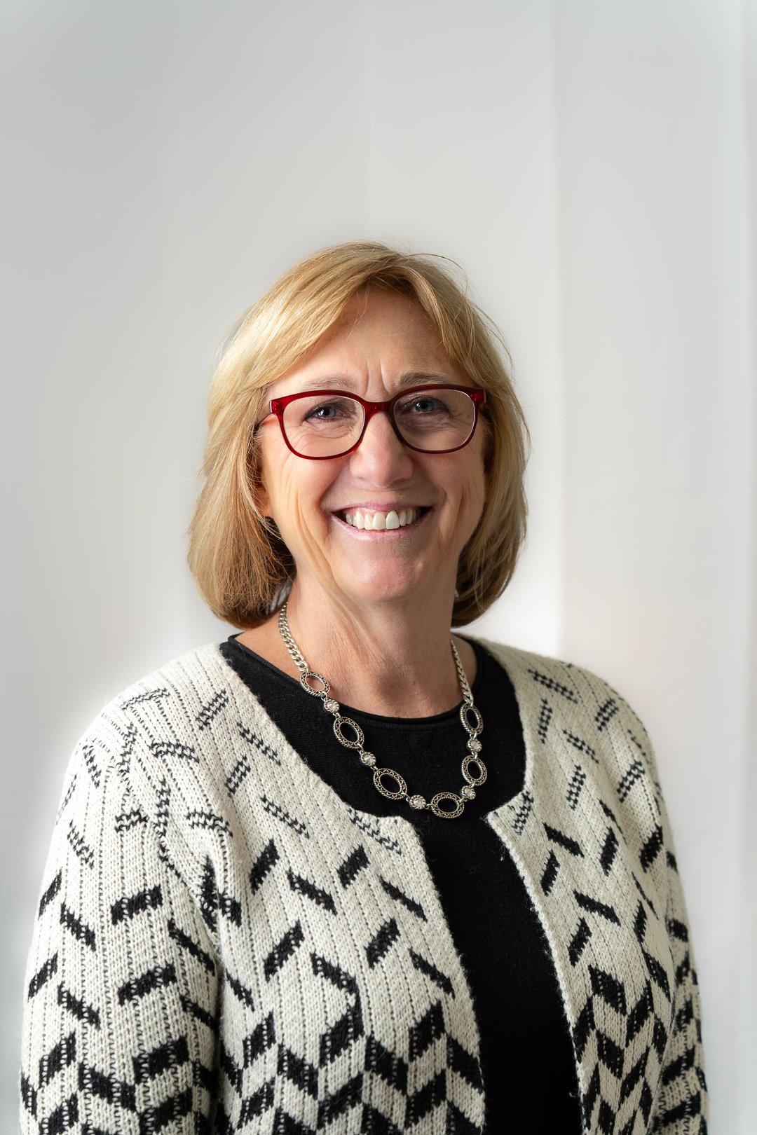 Smiling middle-aged woman with short blonde hair wearing red glasses, a patterned cardigan, and a silver necklace.