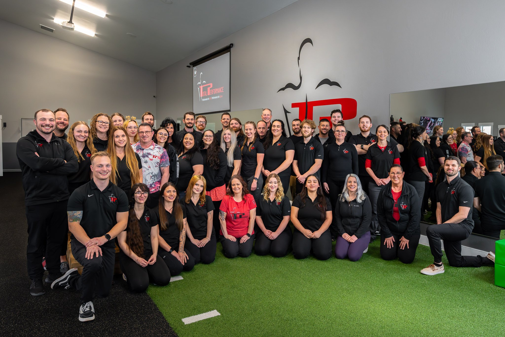 Large group of people posing indoors on artificial grass, many wearing black shirts with a red and white logo.
