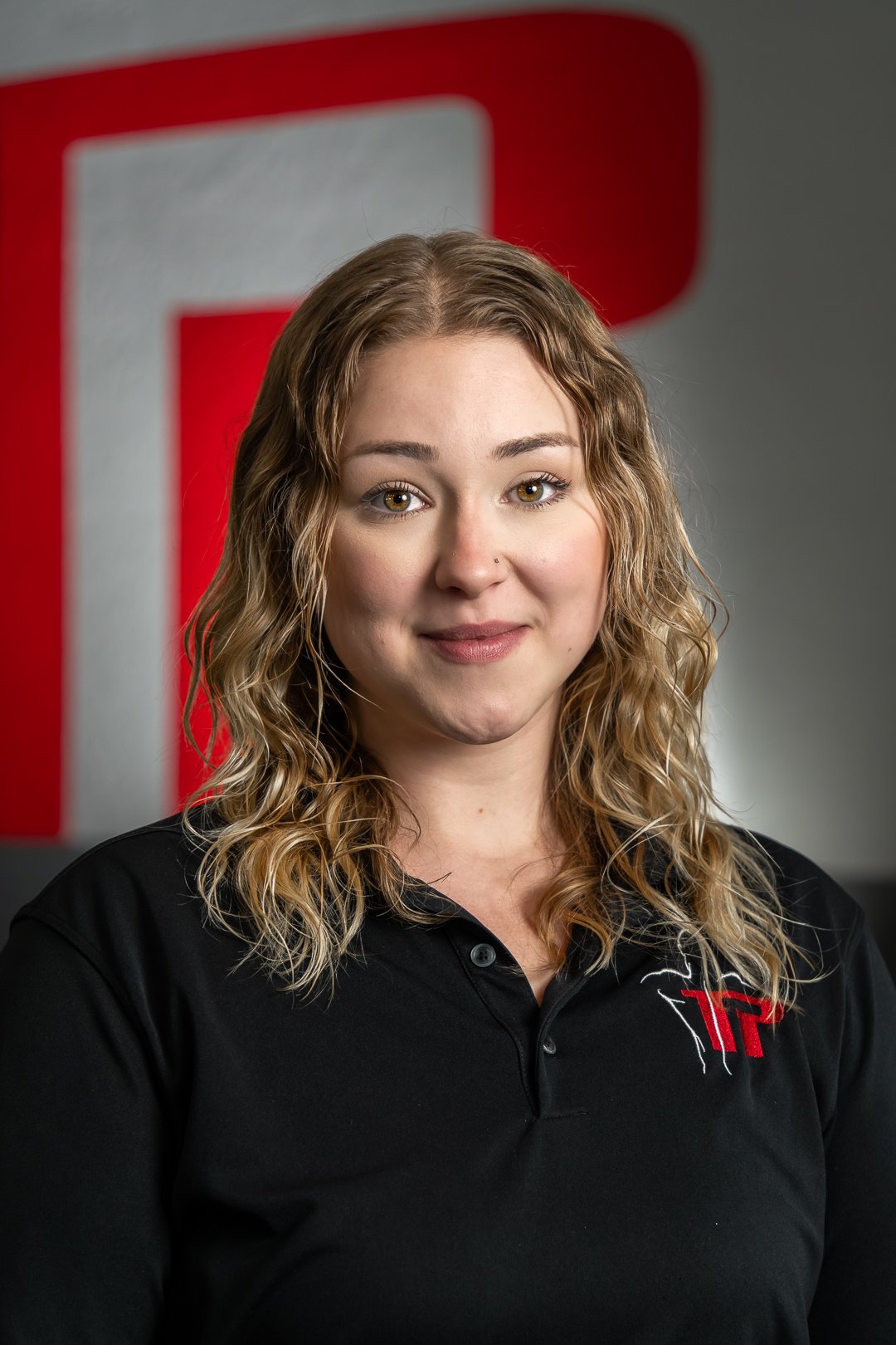 Smiling young woman with wavy blonde hair wearing a black polo shirt with a red and white logo, against a gray background with a red and white geometric design.
