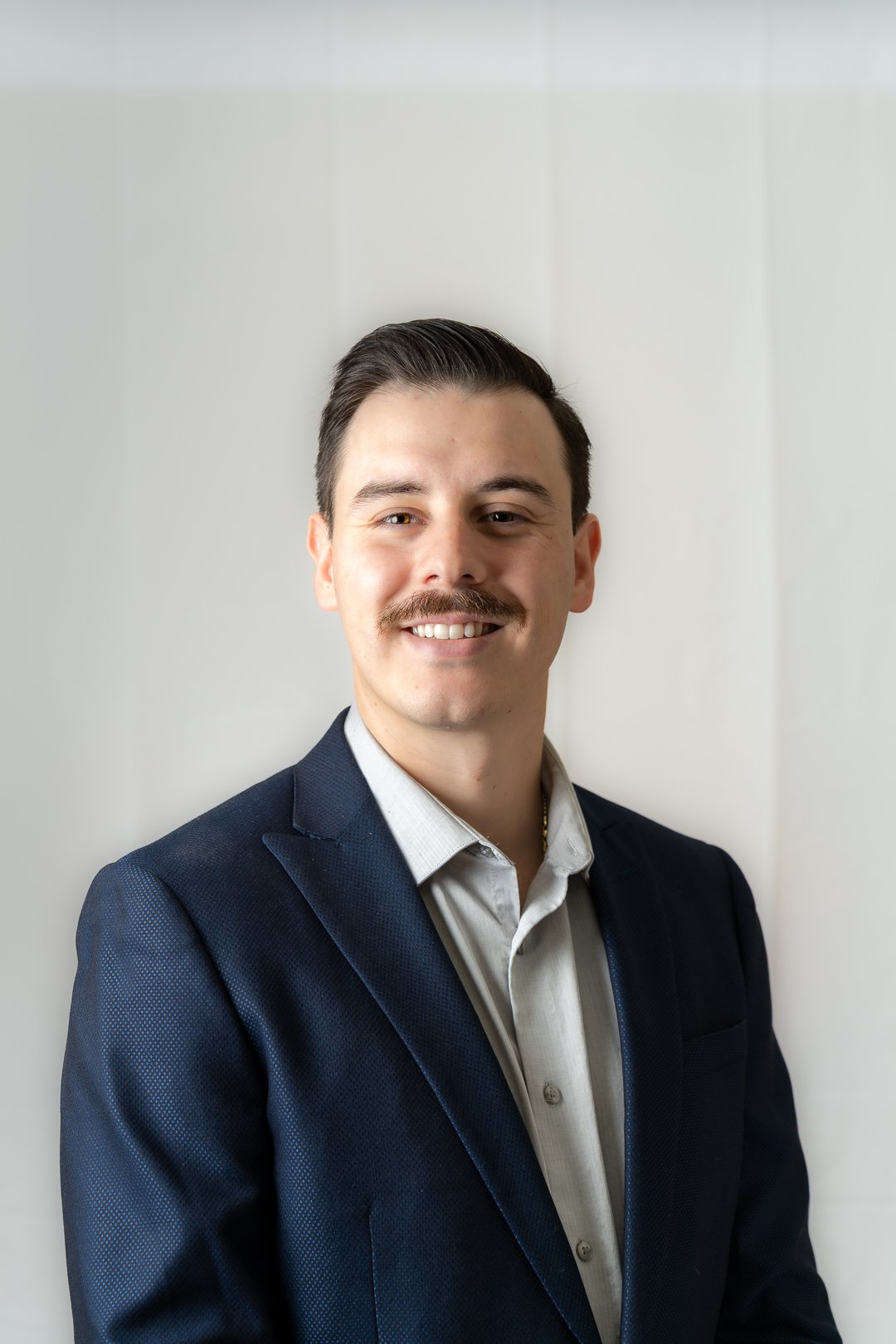 Smiling man with a mustache wearing a navy blazer and light gray shirt against a plain light background.