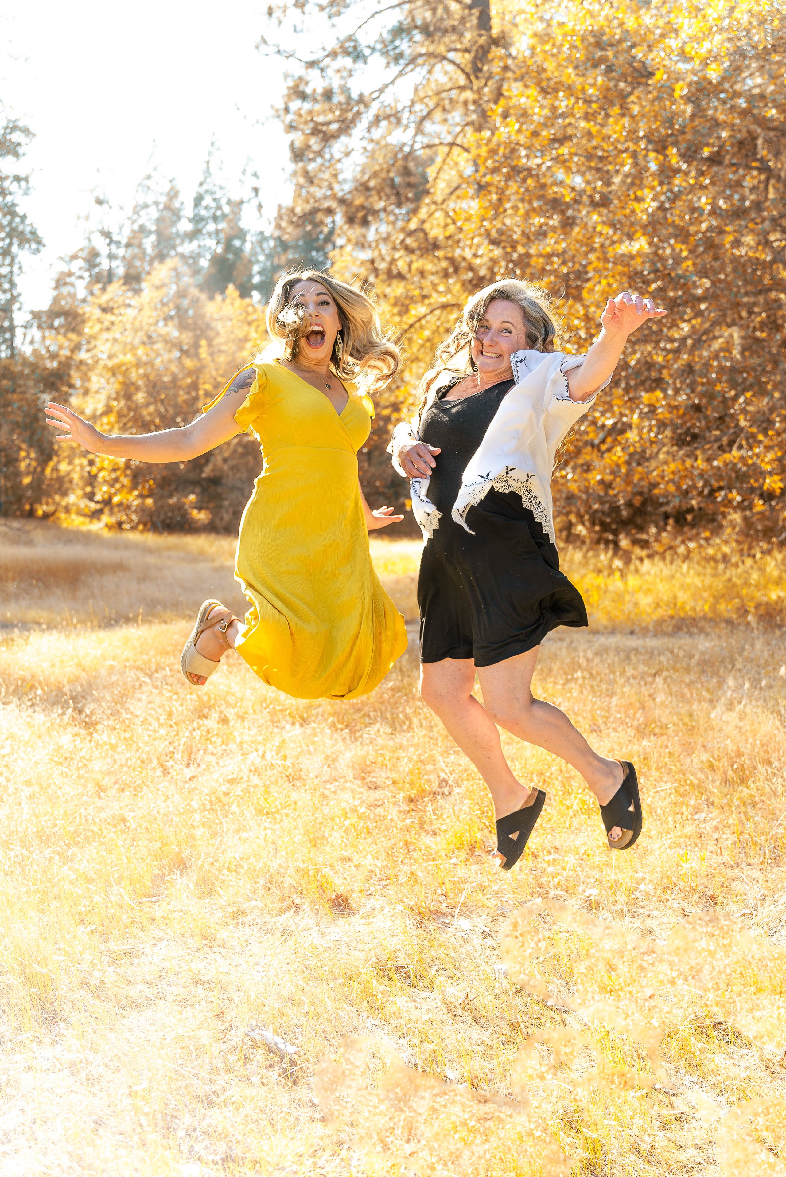 Two women joyfully jumping in a sunlit field with autumn trees in the background.