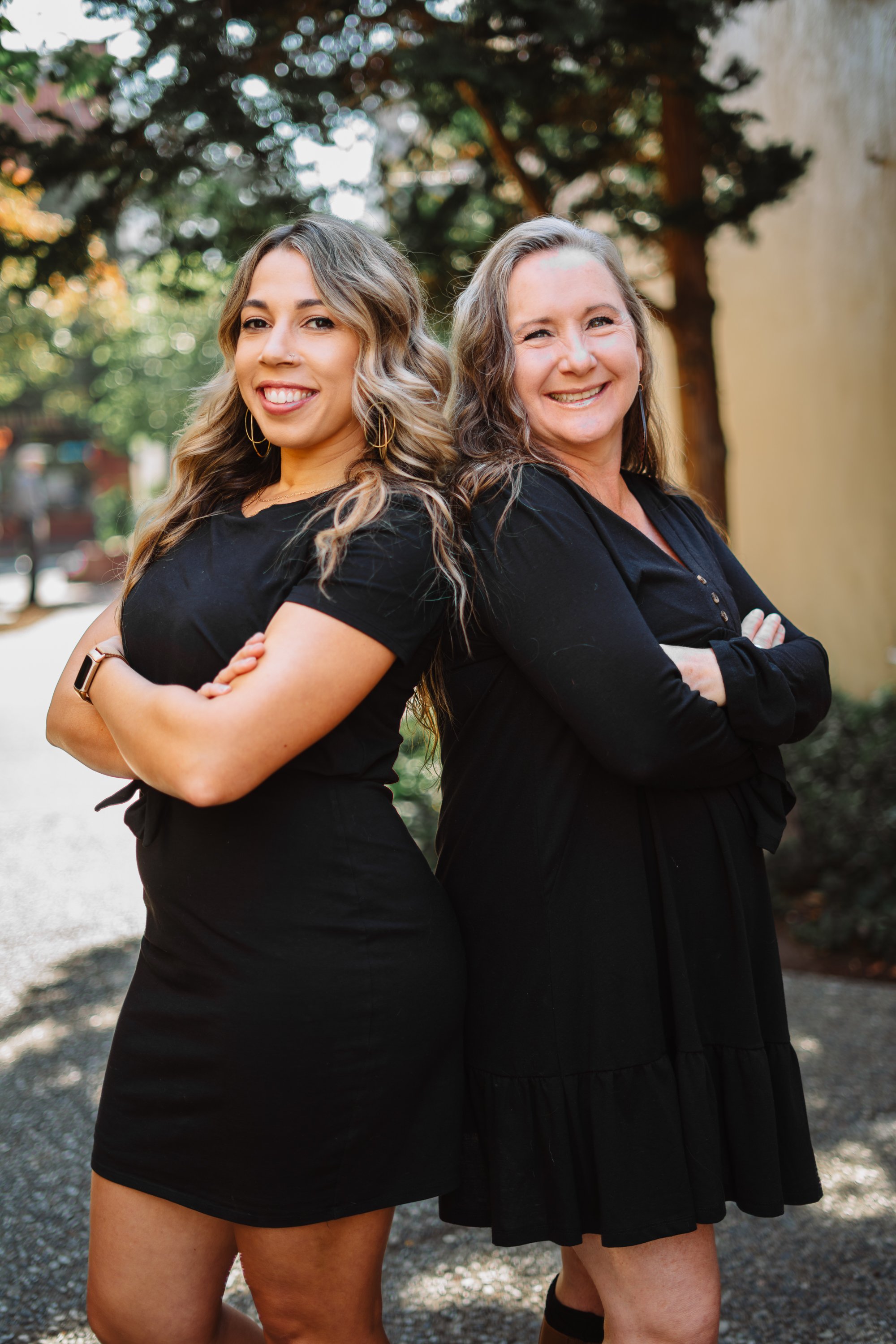 Two women wearing black dresses standing back to back outdoors with arms crossed, smiling at the camera.