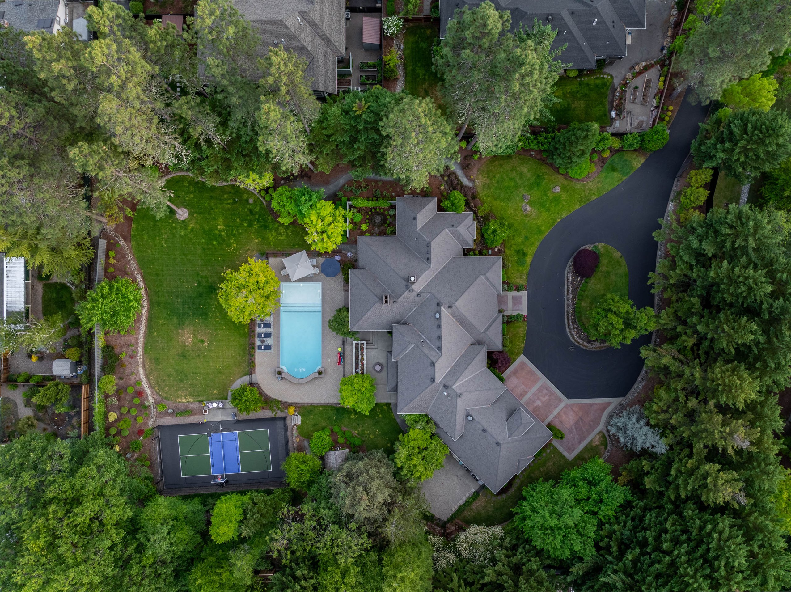 Aerial view of a large house surrounded by trees, a swimming pool, a tennis court, and a curved driveway on a lush green property.