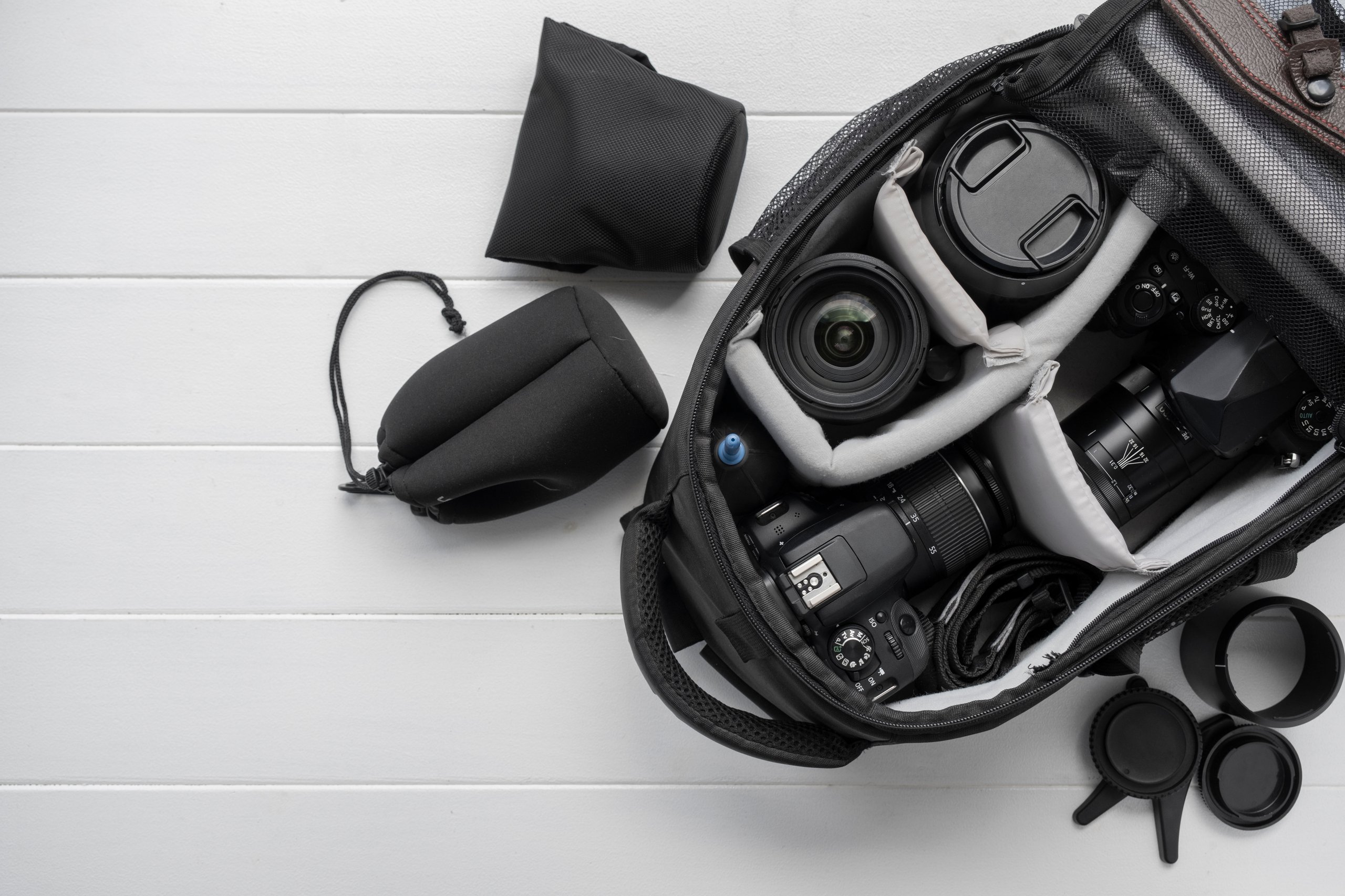 Open black camera bag on white wooden surface containing two DSLR cameras with lenses, extra lens covers, and padded compartments.