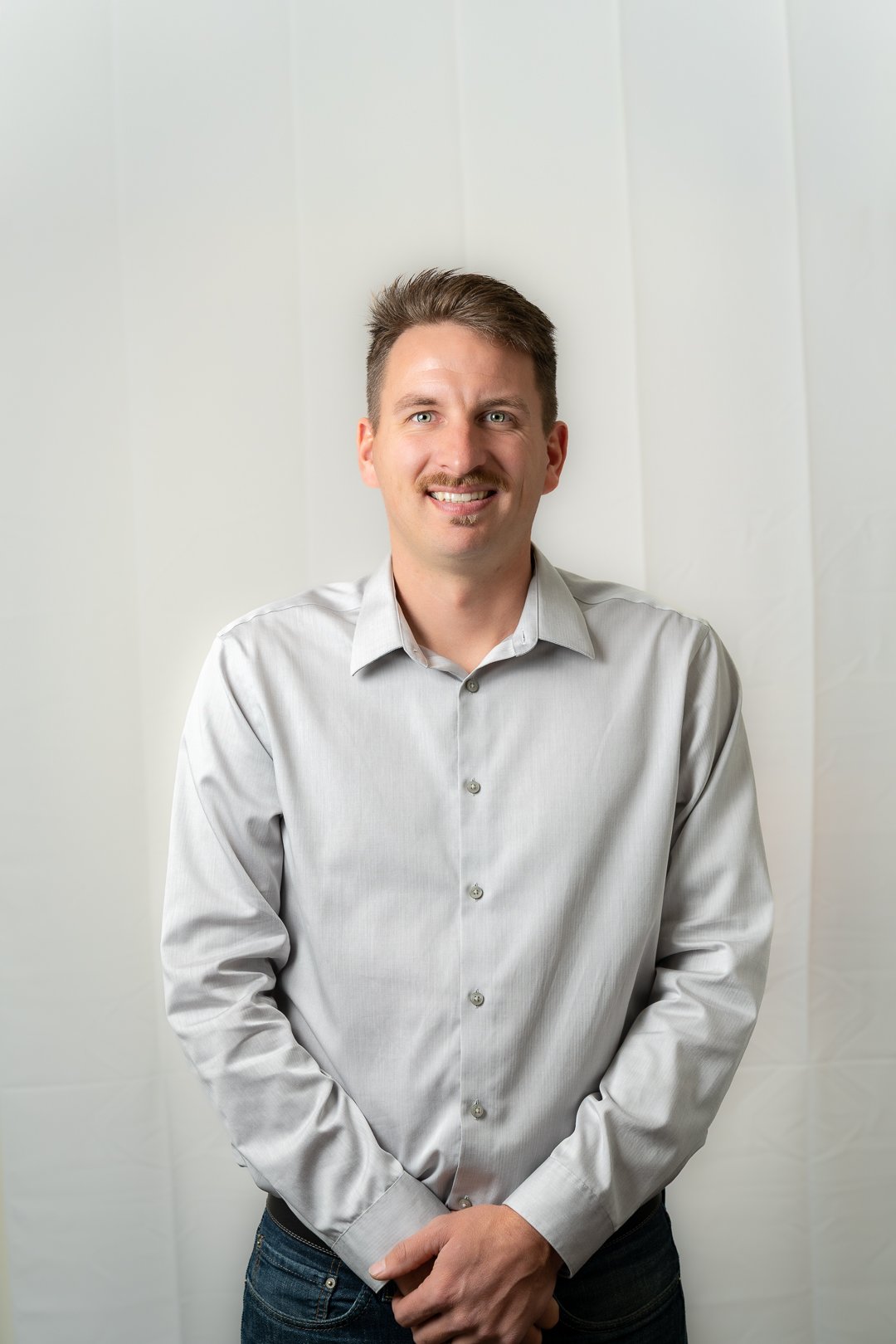 Smiling man with a mustache wearing a light gray button-up shirt and jeans standing against a plain light background.