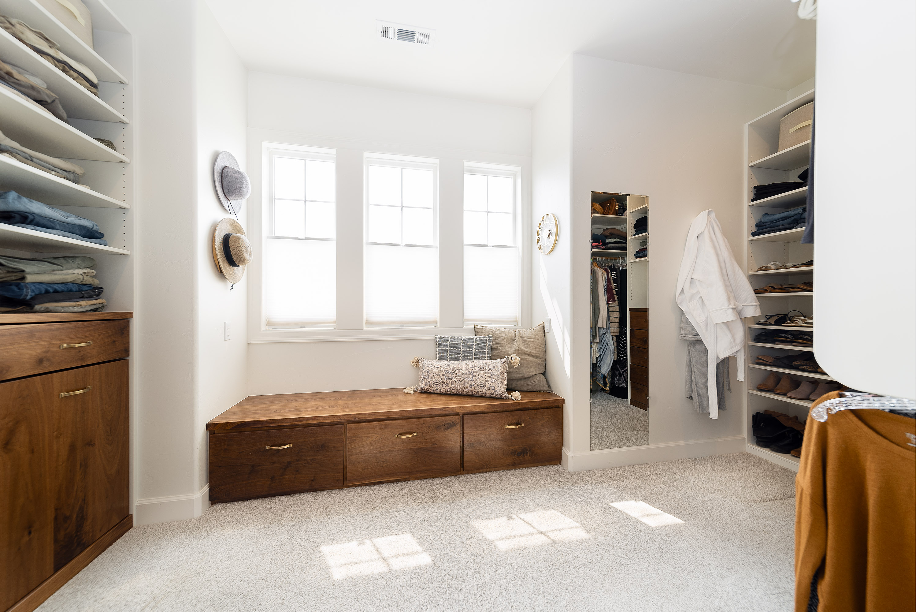 Bright walk-in closet with white walls, wooden drawers, shelves with folded clothes and shoes, a wall-mounted mirror, and a bench with cushions under three windows.