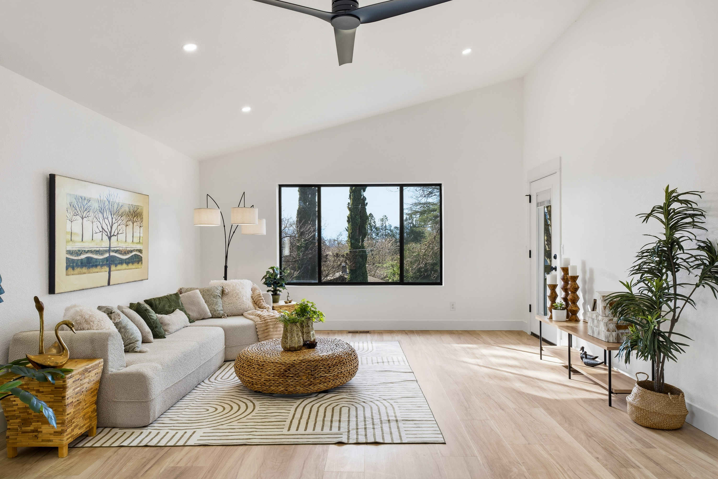 Bright living room with a beige sectional sofa, green and beige pillows, a round wicker coffee table on a patterned rug, large window, and modern decor with plants and wooden accents.