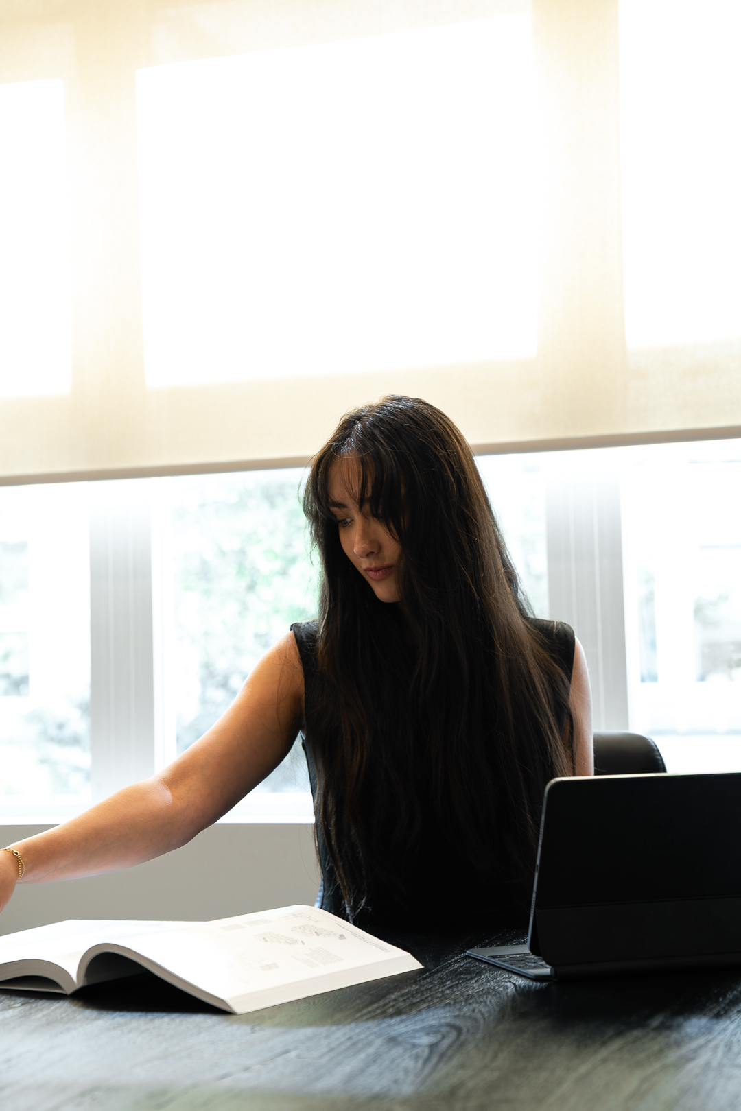 Woman with long dark hair sitting at a table with a book and a tablet, looking down thoughtfully.