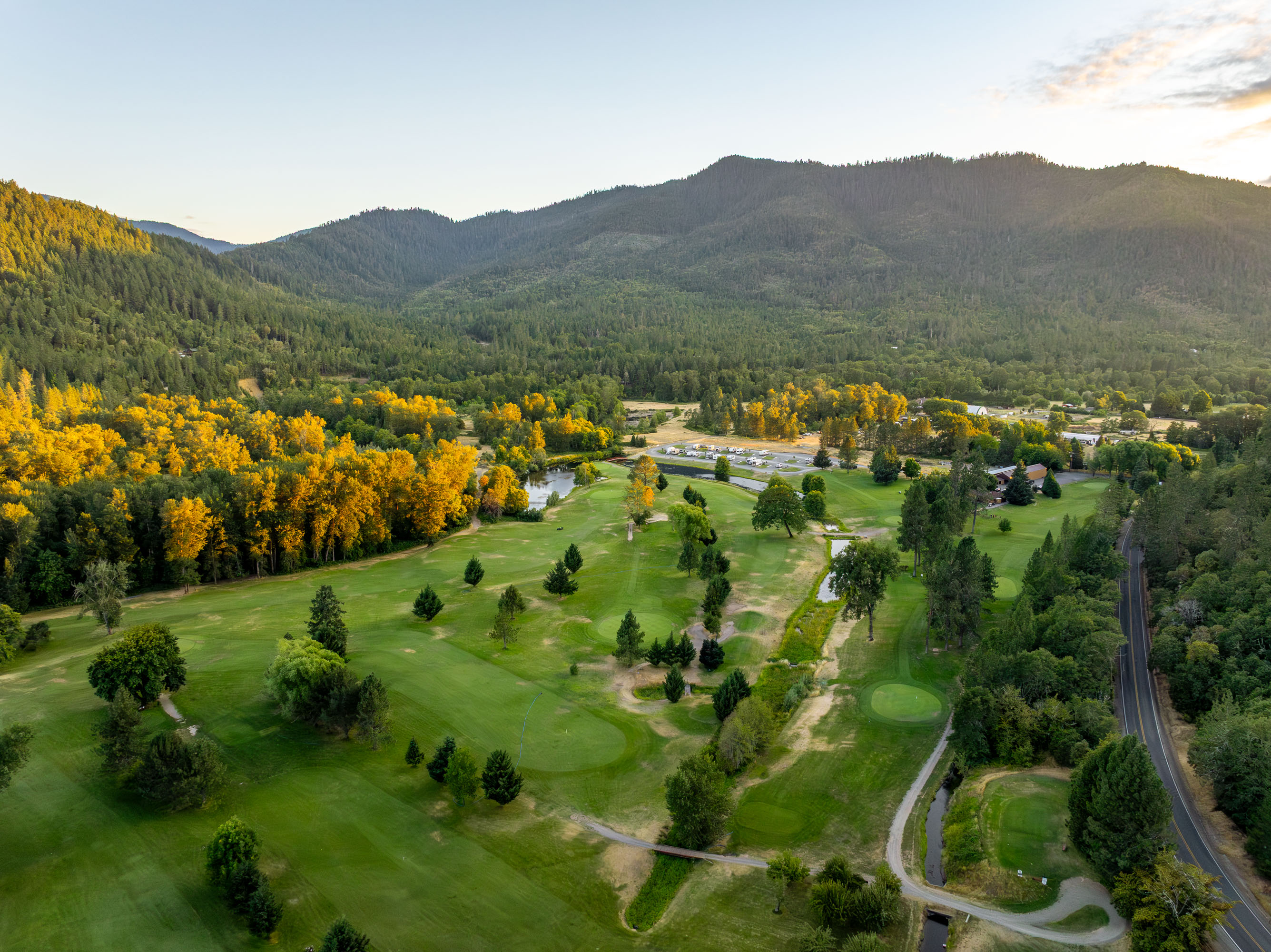 Aerial view of a golf course surrounded by lush trees with yellow autumn foliage, a small pond, and mountains in the background.