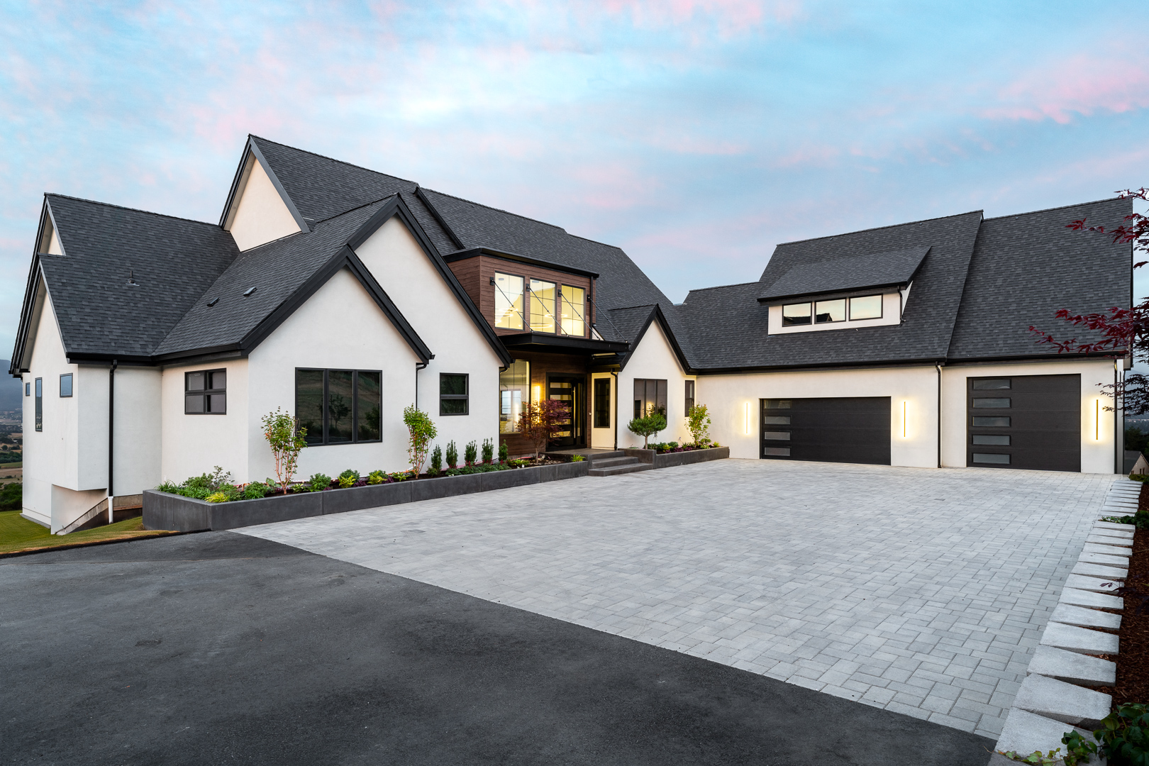 Modern two-story house with white walls, dark gray roof, large driveway, and illuminated exterior lights at dusk.