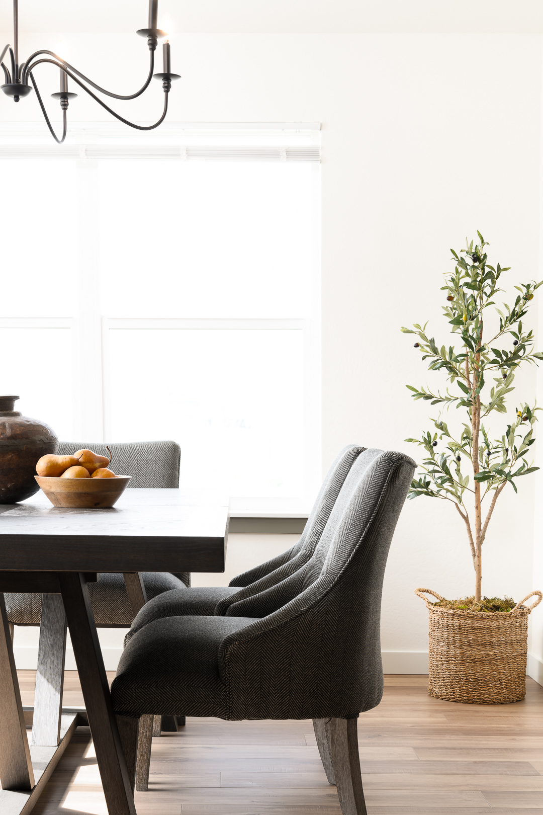 Modern dining area with dark upholstered chairs, a wooden table with a bowl of pears, and a potted olive tree in a woven basket by a sunlit window.