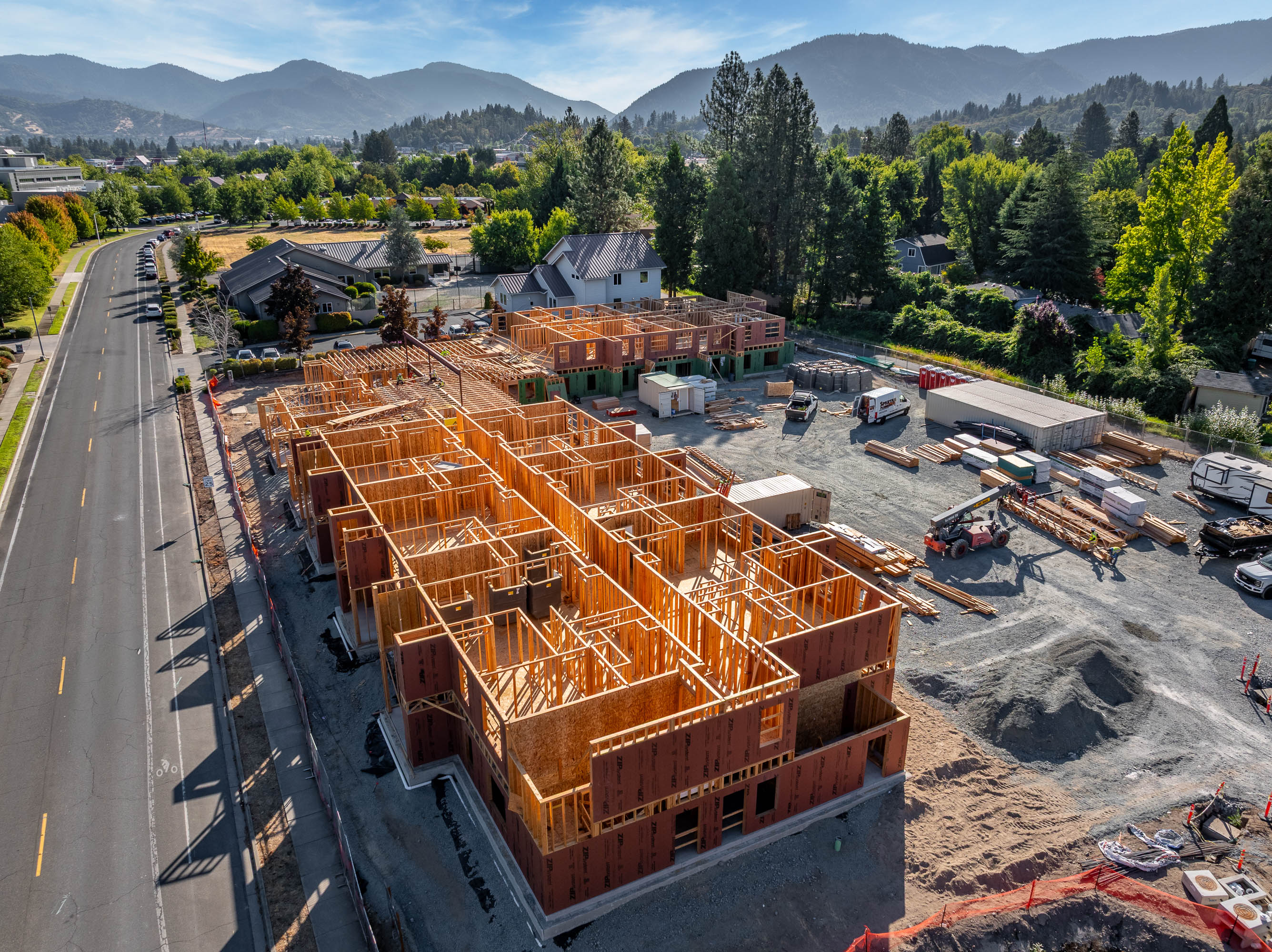Aerial view of a large wood-framed building under construction beside a street, with vehicles, construction materials, and mountains in the background.