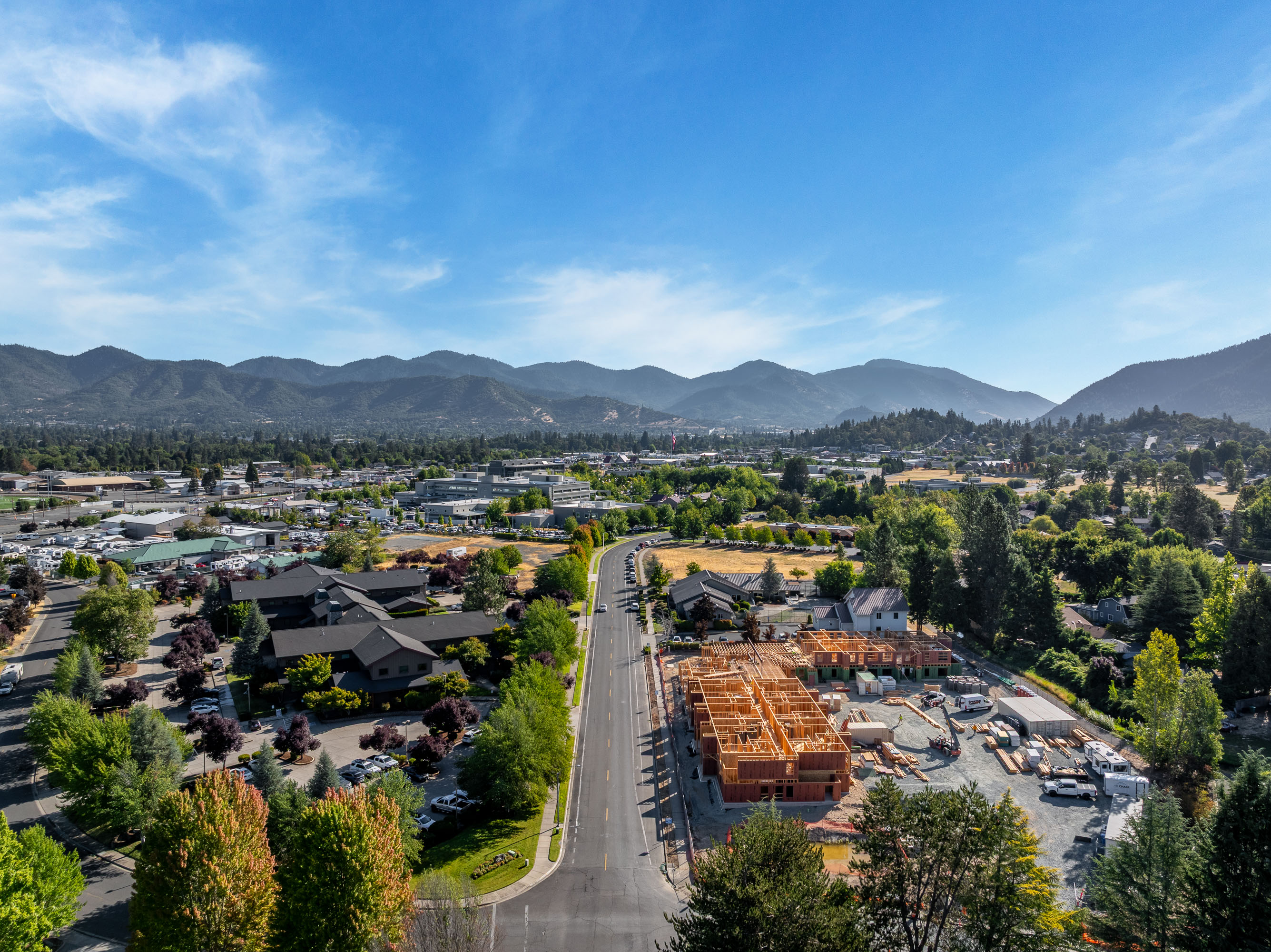 Aerial view of a suburban area with a road running through, houses, buildings, a construction site with wooden frames, surrounded by trees and mountains under a blue sky.