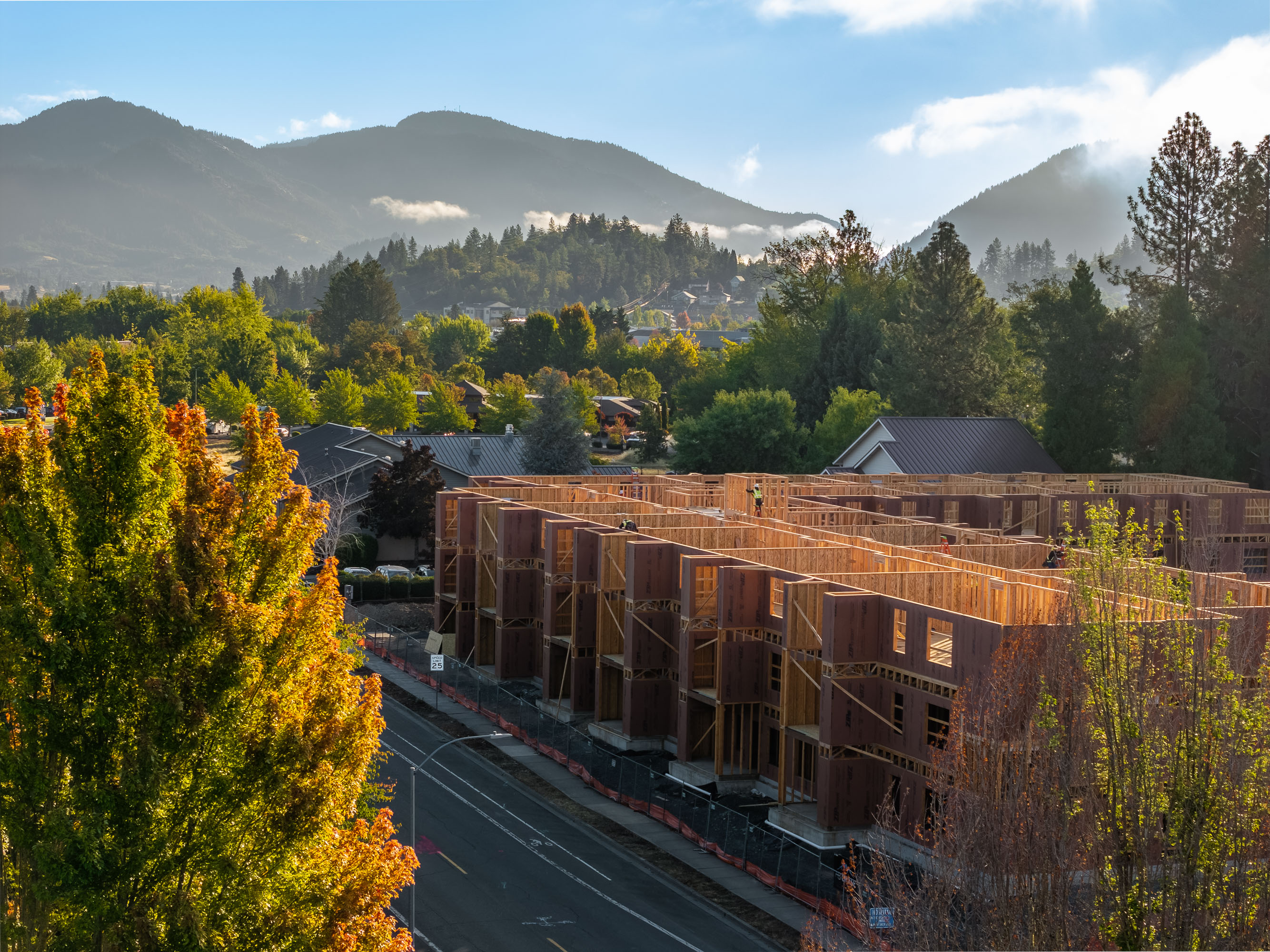 Construction site of wooden framed buildings beside a road with trees and mountains in the background under a clear sky.