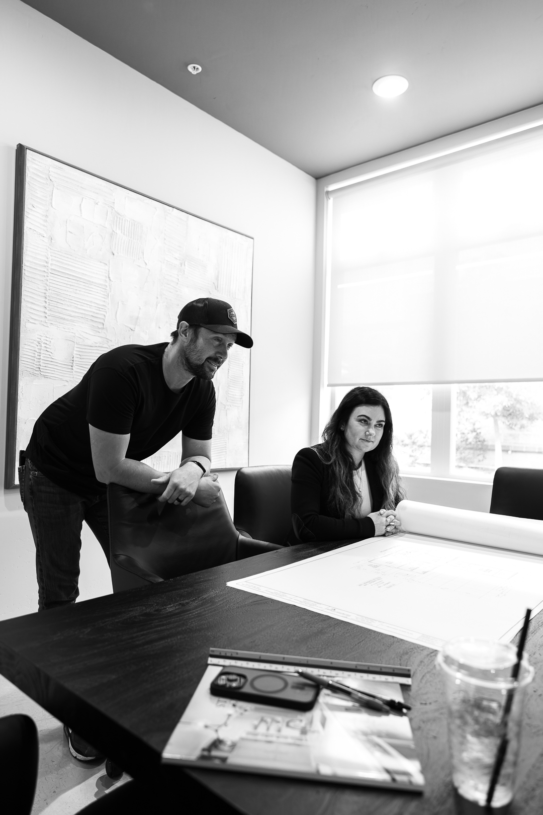Man leaning on chair and woman sitting at a table reviewing architectural plans in a bright office.