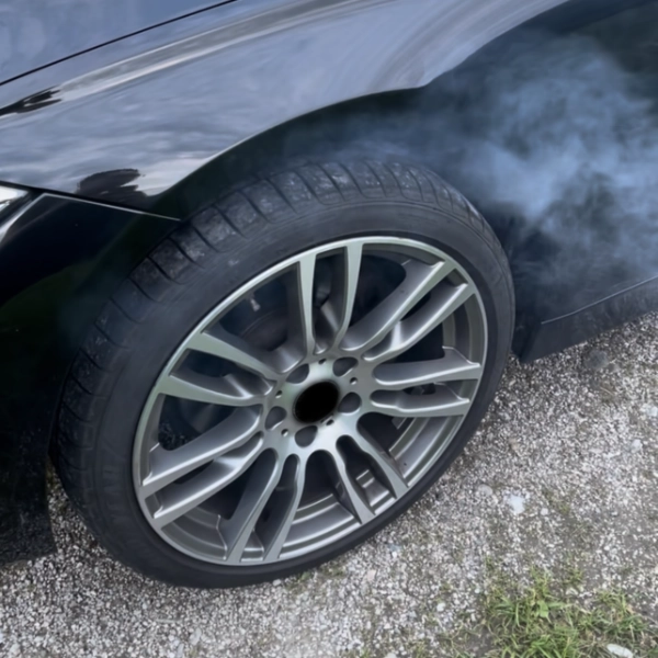 Close-up of a black car's wheel with a alloy rim and smoke rising from the tire on a gravel surface.