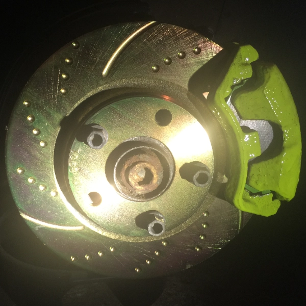 Close-up of a metallic drilled and slotted brake rotor with a bright green brake caliper attached.