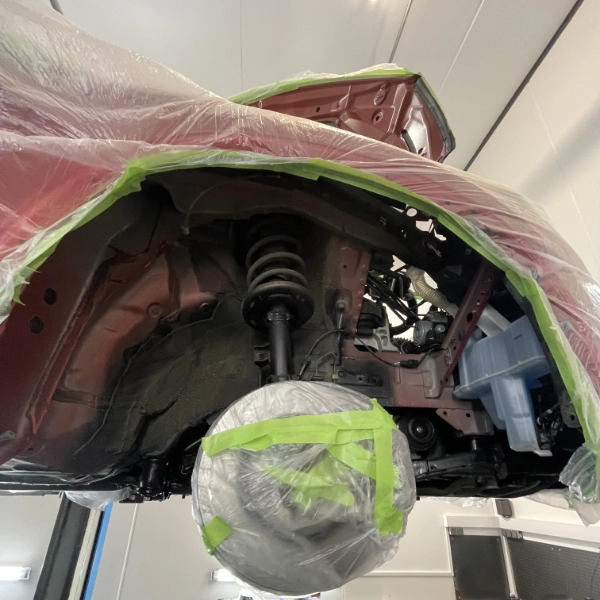 Car lifted with wheel removed, suspension and brake components covered with protective plastic wrap and tape in a repair workshop.
