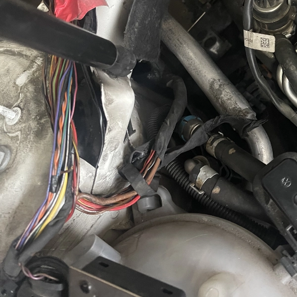 Close-up of a car engine compartment showing a bundle of colorful electrical wires wrapped with black tape, various hoses, and mechanical components.
