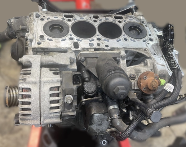 Disassembled car engine block showing four cylinder chambers and attached alternator and fuel filter components.