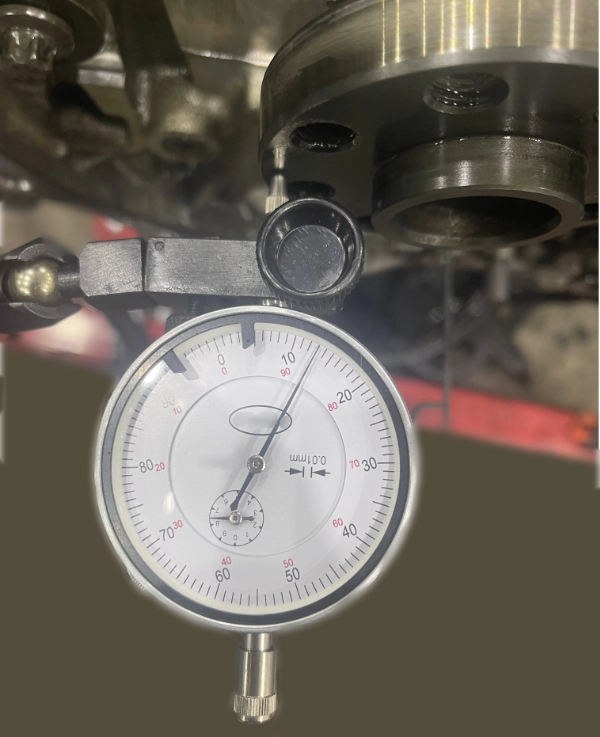 Dial indicator gauge measuring axial play on a metallic mechanical part with a 0.01 mm scale.